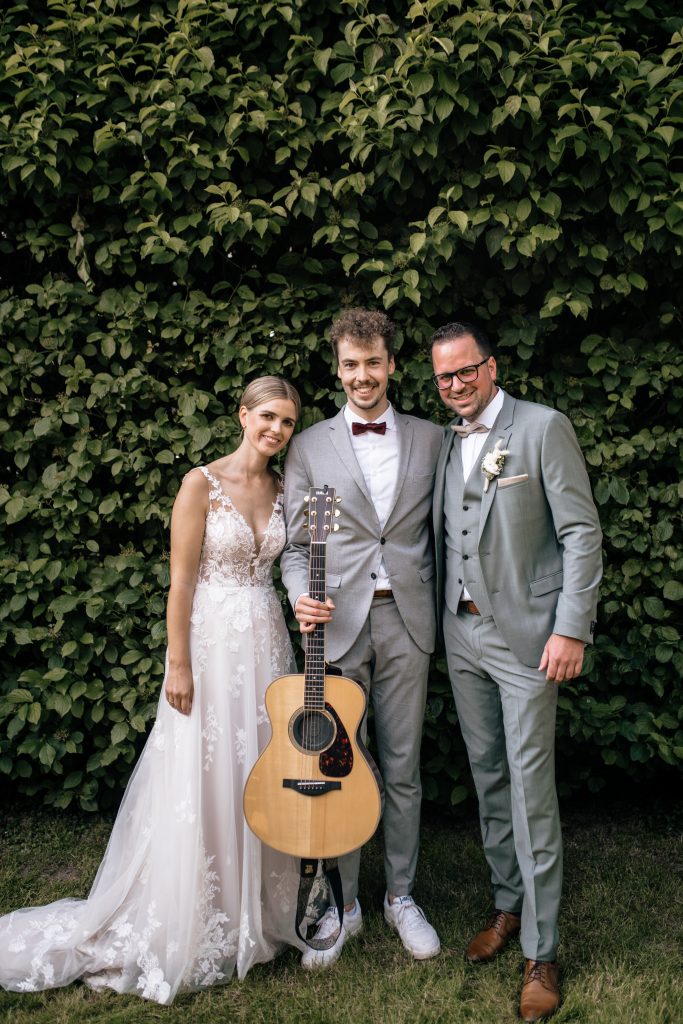 ochzeitssänger aus Köln mit Akustikgitarre steht lächelnd neben einem frisch vermählten Brautpaar vor einer grünen Hecke.